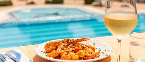 A dining table with a plate of seafood, a glass of white wine, and a pool in the background.