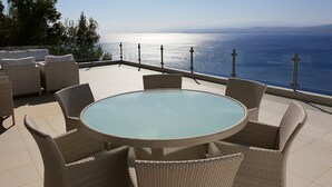 A round glass table with wicker chairs on a terrace overlooking a body of water.