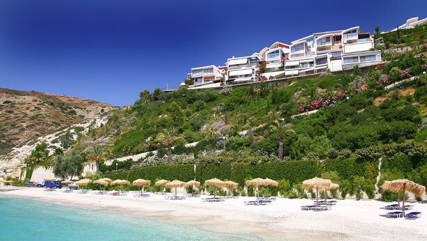 A coastal hotel with multiple buildings on a hillside, a beach with umbrellas, and clear turquoise waters.