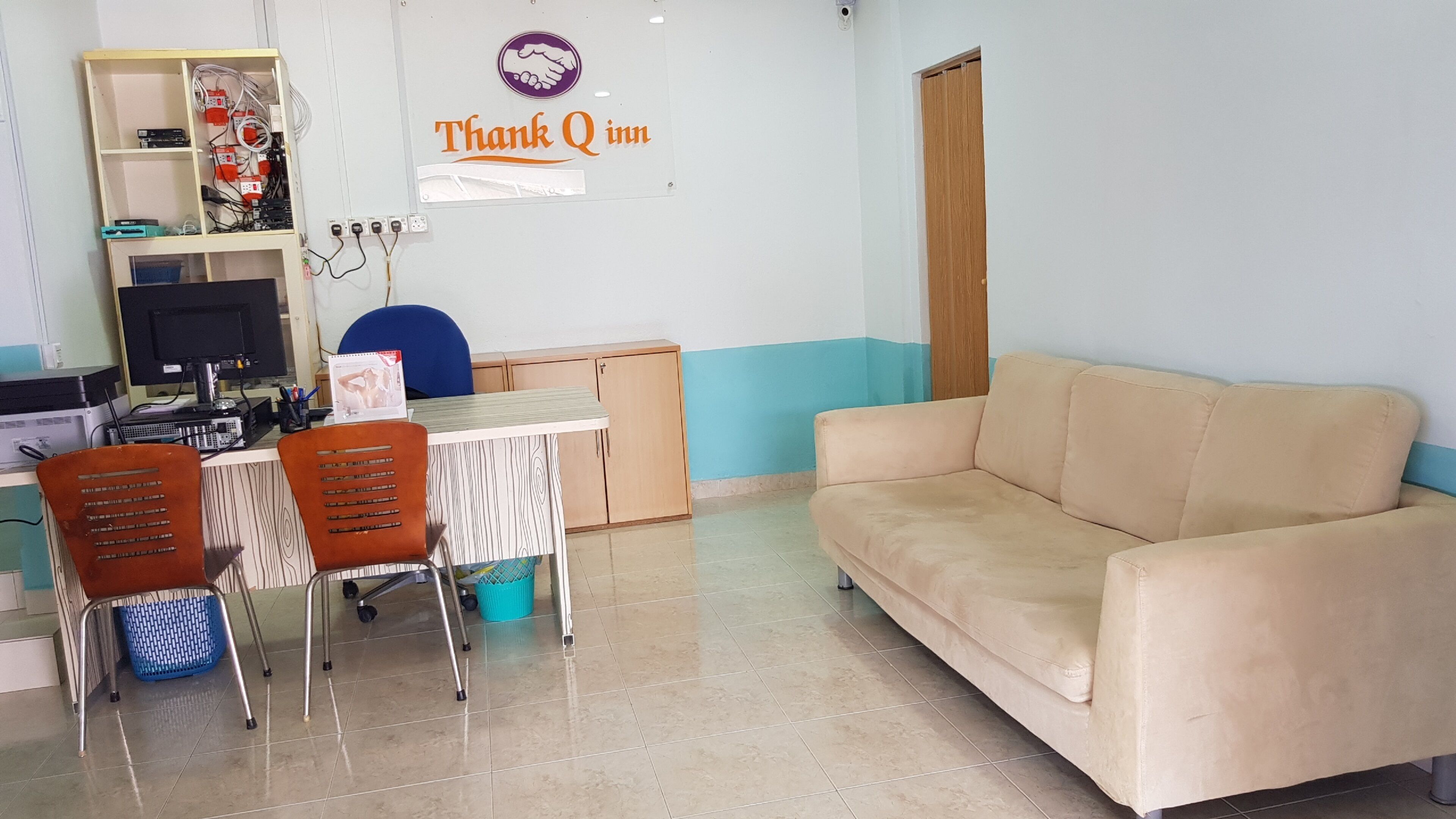 A room with a beige sofa, a desk with a computer, chairs, and a blue chair. There is a sign on the wall that reads "Thank Q inn".