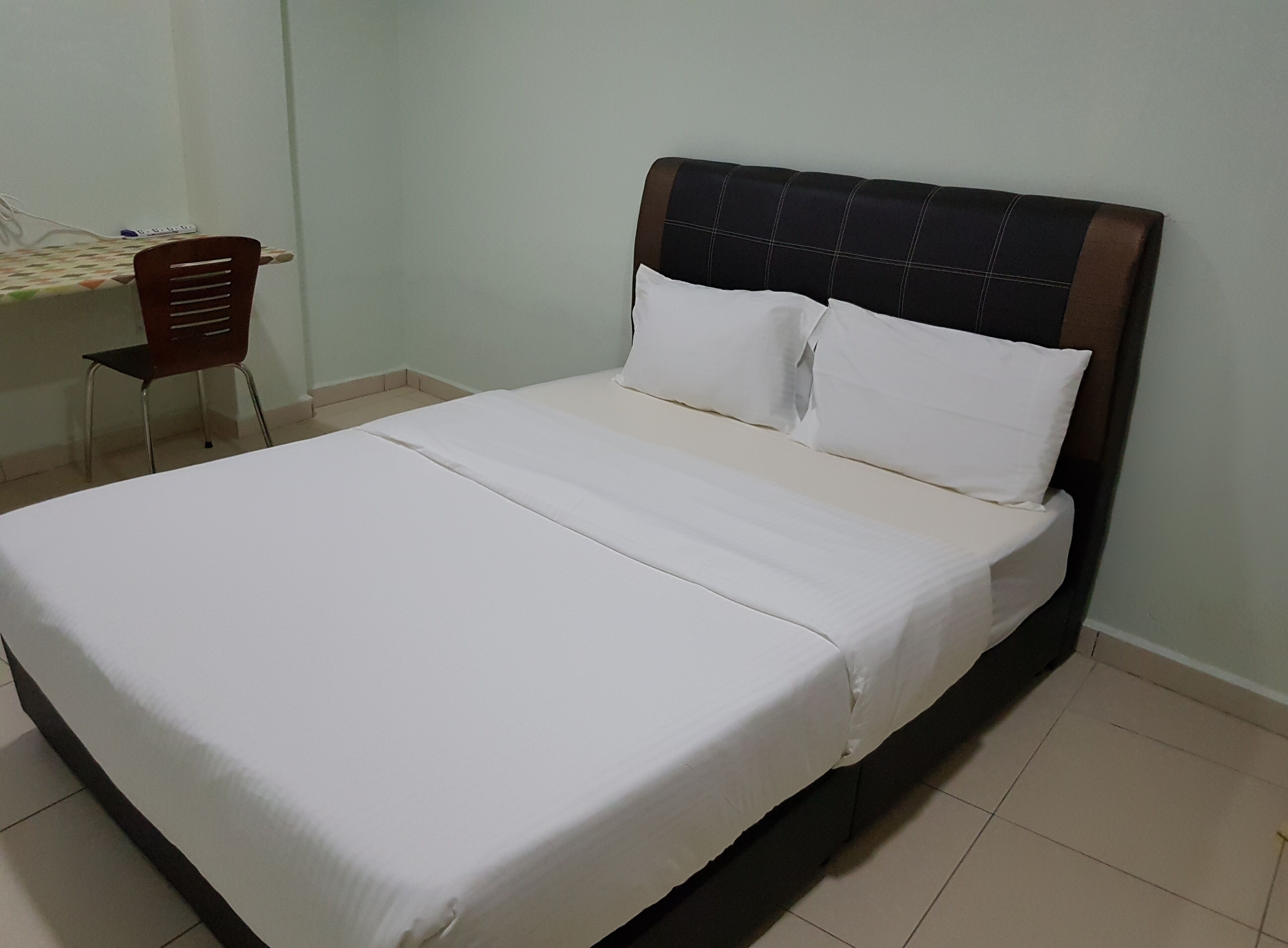 A neatly made bed with white linens and a dark headboard in a room with a tiled floor and a desk with a chair.