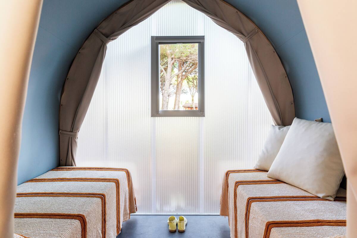 Ein Zimmer mit zwei Betten, einem kleinen Tisch mit Schuhen und einem Fenster mit Blick auf Bäume.