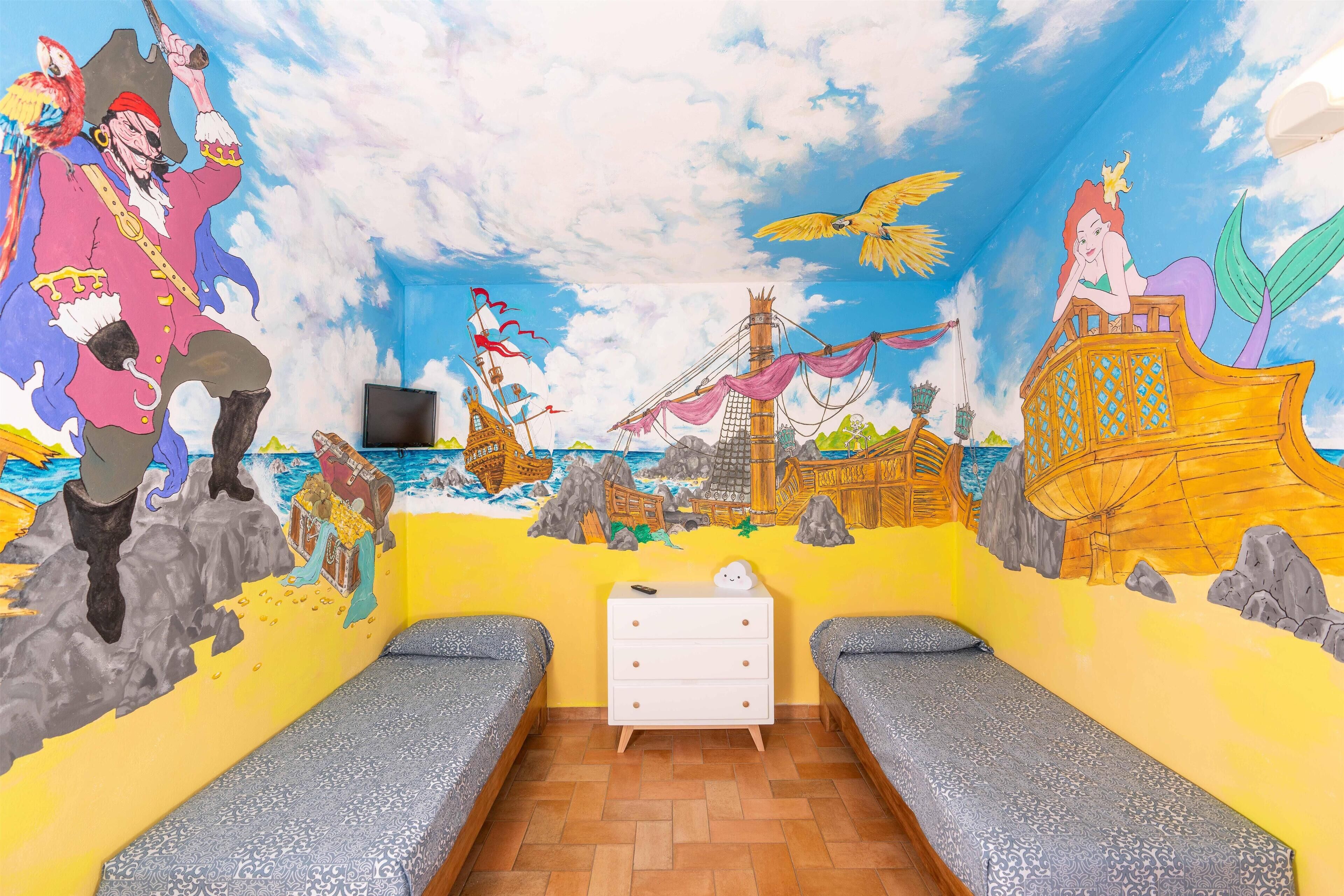 A room with a mural of a pirate and mermaid, two beds, a white dresser, and a TV on a stand.