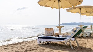 Una playa con tumbonas y sombrillas, una mesa con una toalla y un sombrero, y vistas al mar y a las montañas.