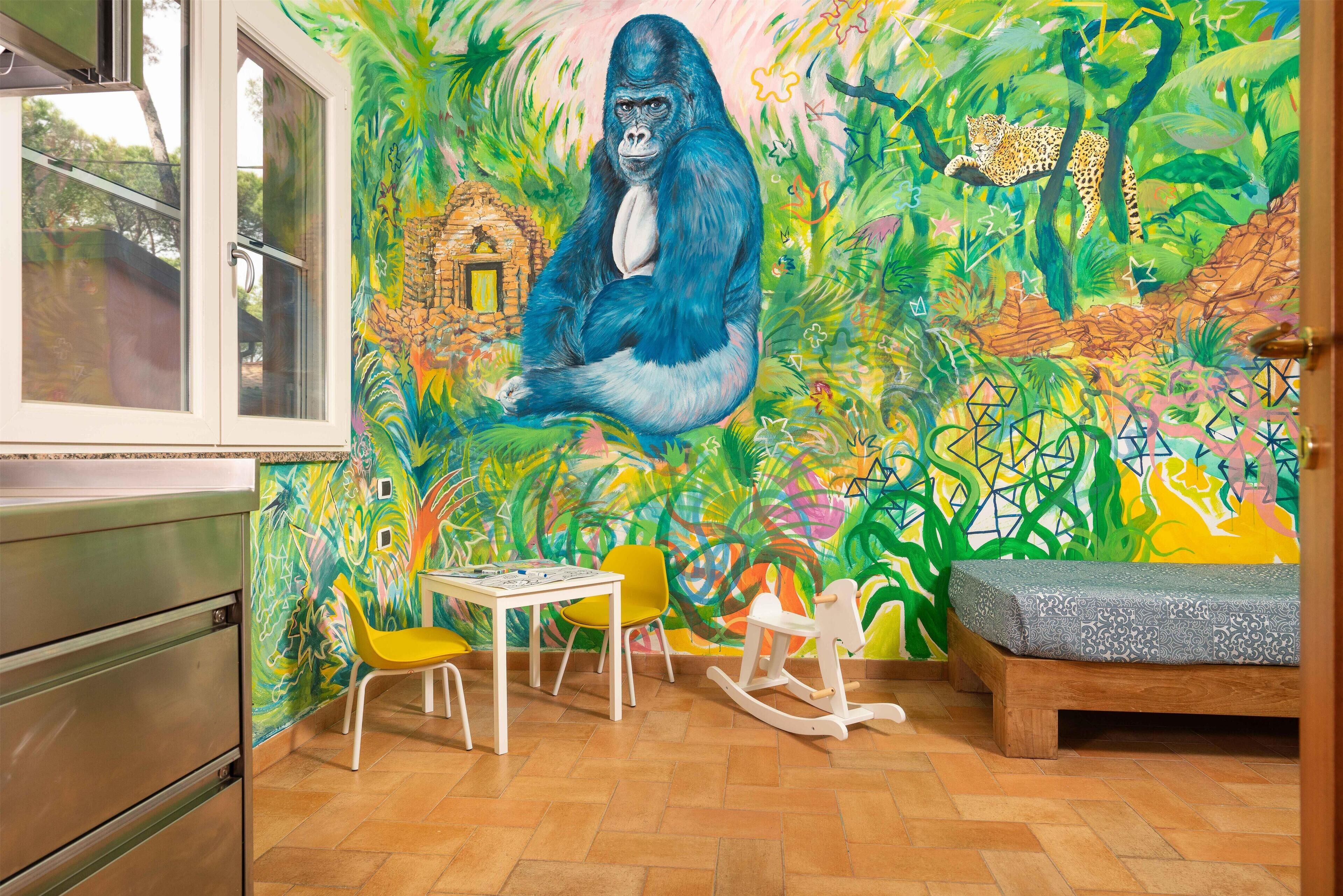 A room with a vibrant mural, a wooden bed, a small table with chairs, and a kitchen area.