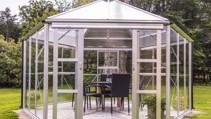 A glass greenhouse with a table and chairs, surrounded by a grassy area and trees. - Pederstrup Bed and Breakfast (Torrig)
