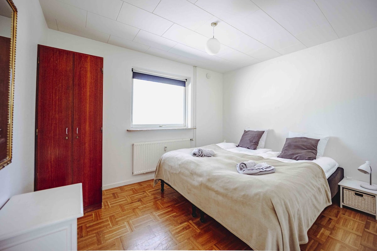 A bedroom with a wooden wardrobe, a bed with pillows, a bedside table with a lamp, and a window with a blind.