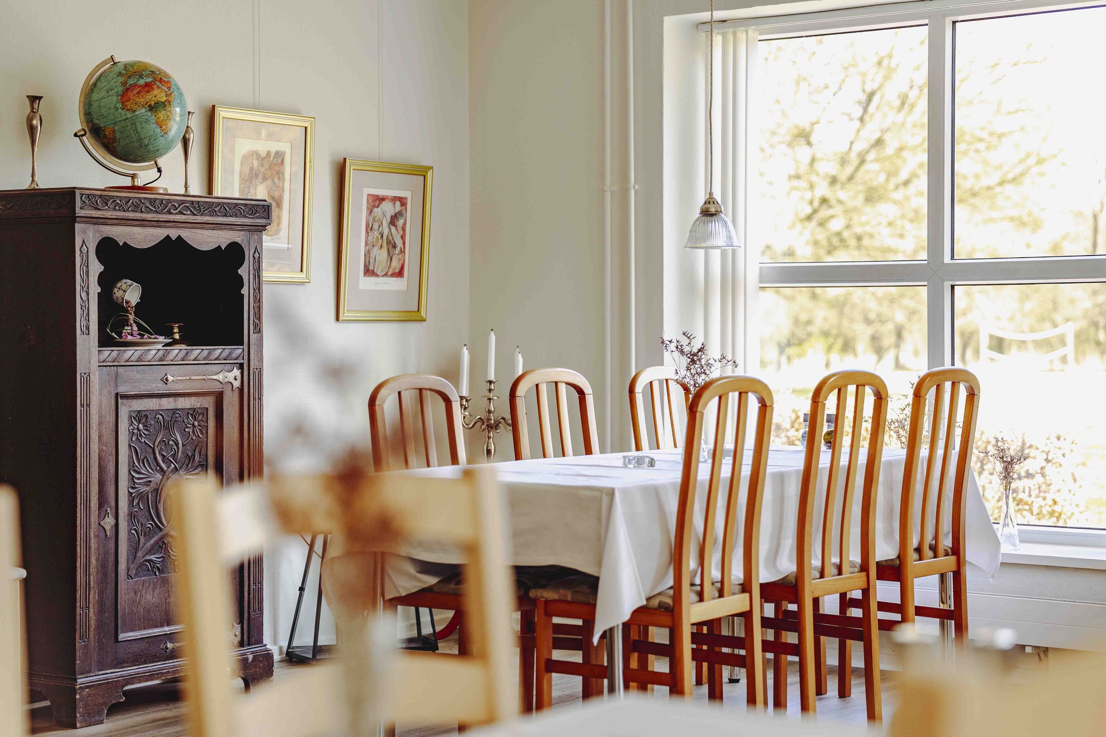A dining area with a table set for four, wooden chairs, a globe on a cabinet, framed pictures on the wall, and a window with a view of trees.