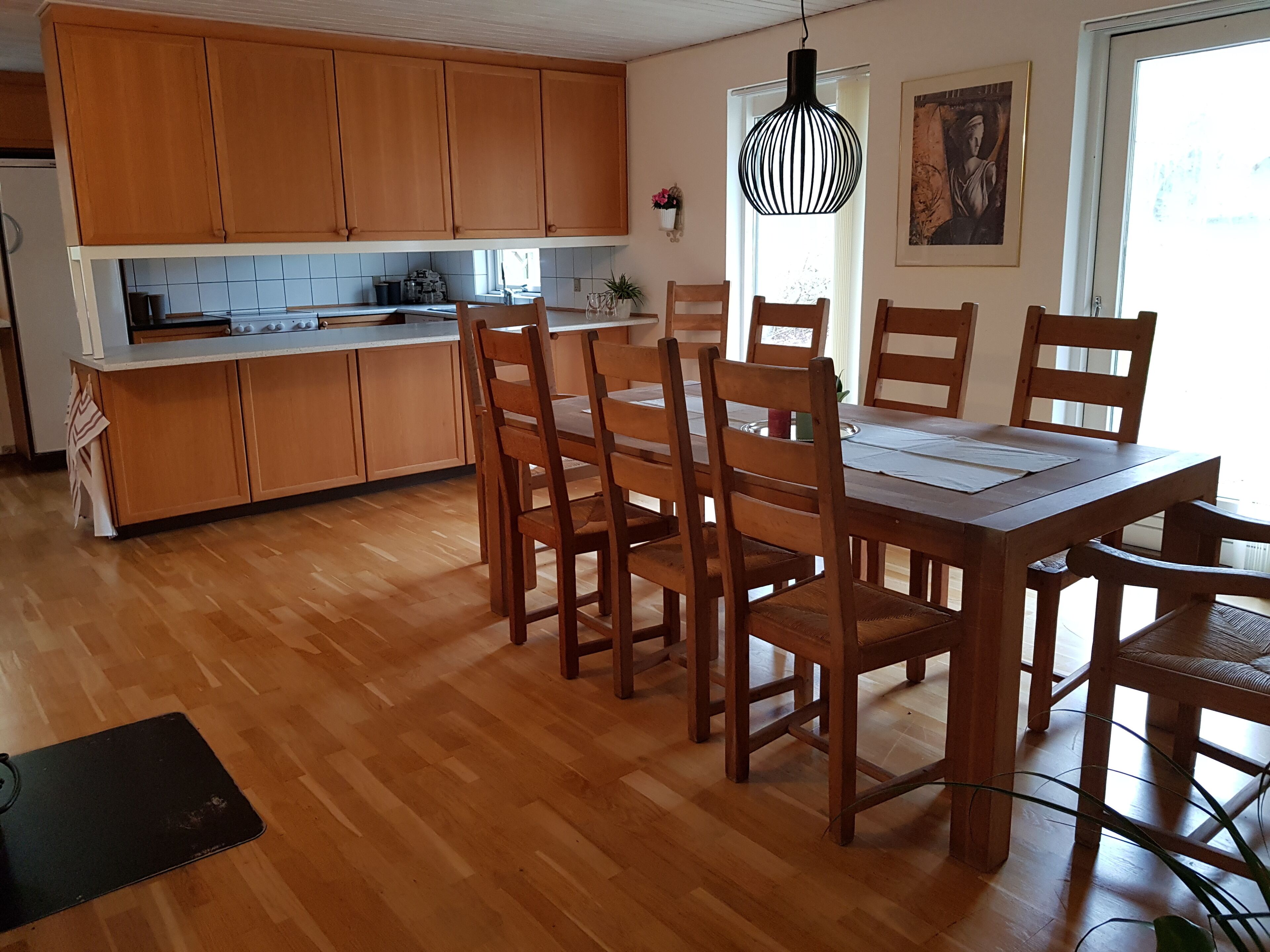 Una cocina con armarios de madera, una mesa de comedor con sillas, una lámpara colgante y una ventana con cortinas.