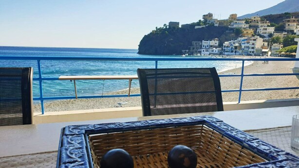 A table with a woven basket and two spherical objects, overlooking a beach with a rocky outcrop and buildings in the background.