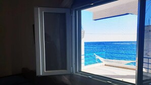 A room with a large window overlooking the ocean, a bed, and a small table with a bag on it.