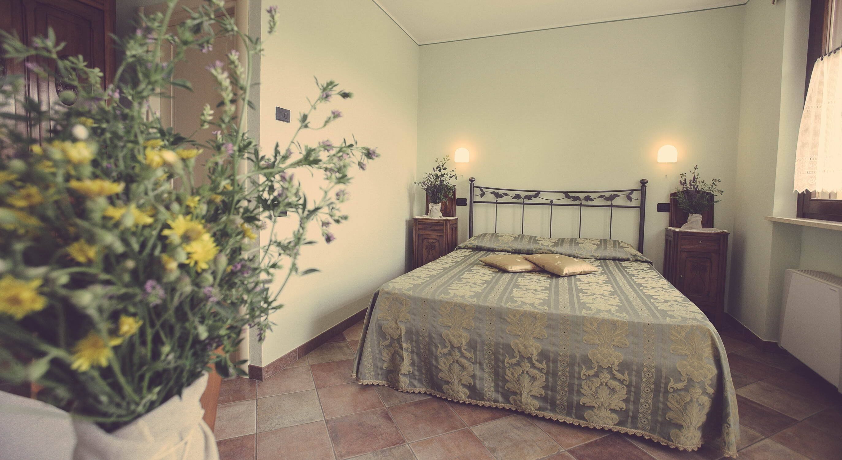 Un dormitorio con una cama, dos mesitas de noche, una planta en maceta y un ventanal con cortinas.