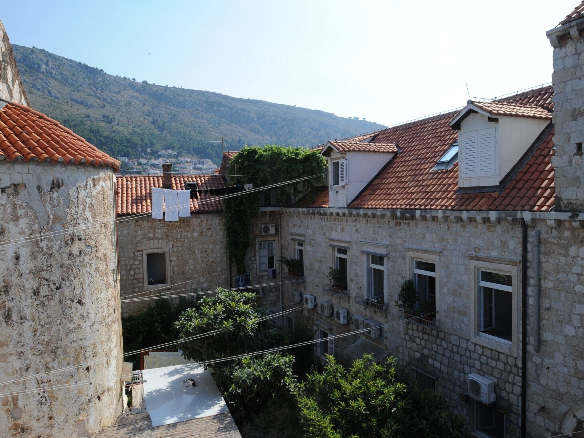 A stone building with a tiled roof and multiple windows, surrounded by greenery and a hilly landscape in the background.
