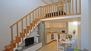 Um apartamento moderno com escada de madeira, cozinha com armários brancos, área de jantar com mesa e cadeiras, e sofá cinza com almofadas verdes.