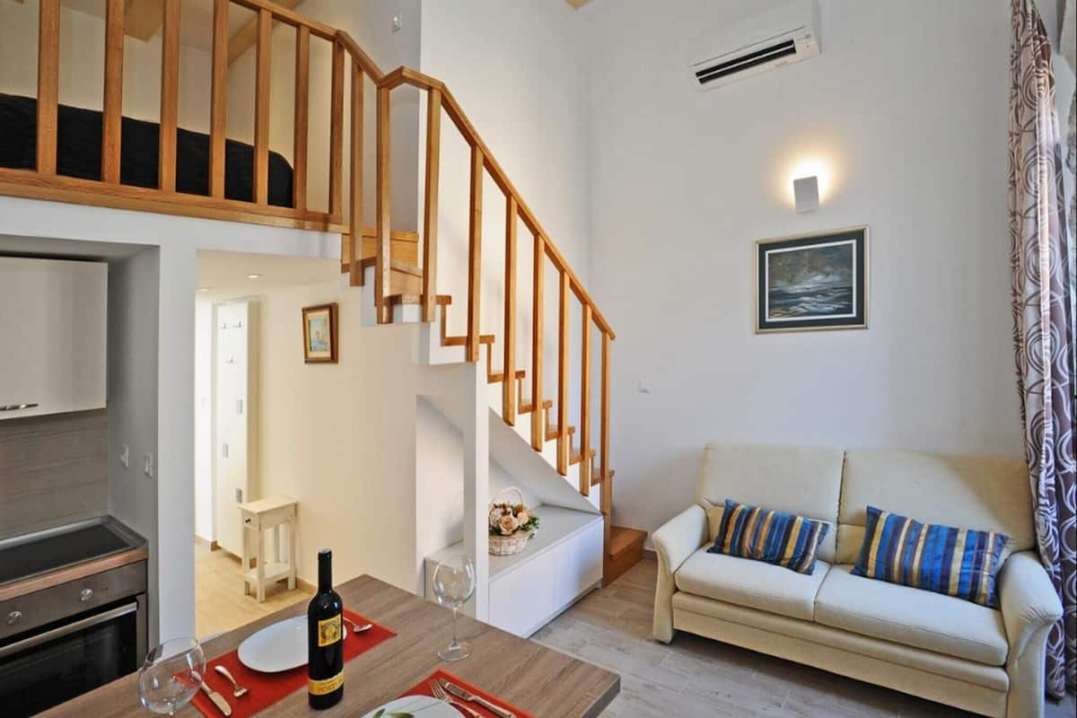Un salon moderne avec un escalier en bois, un canapé, un coin repas avec une table dressée pour deux personnes, et une cuisine visible au fond.