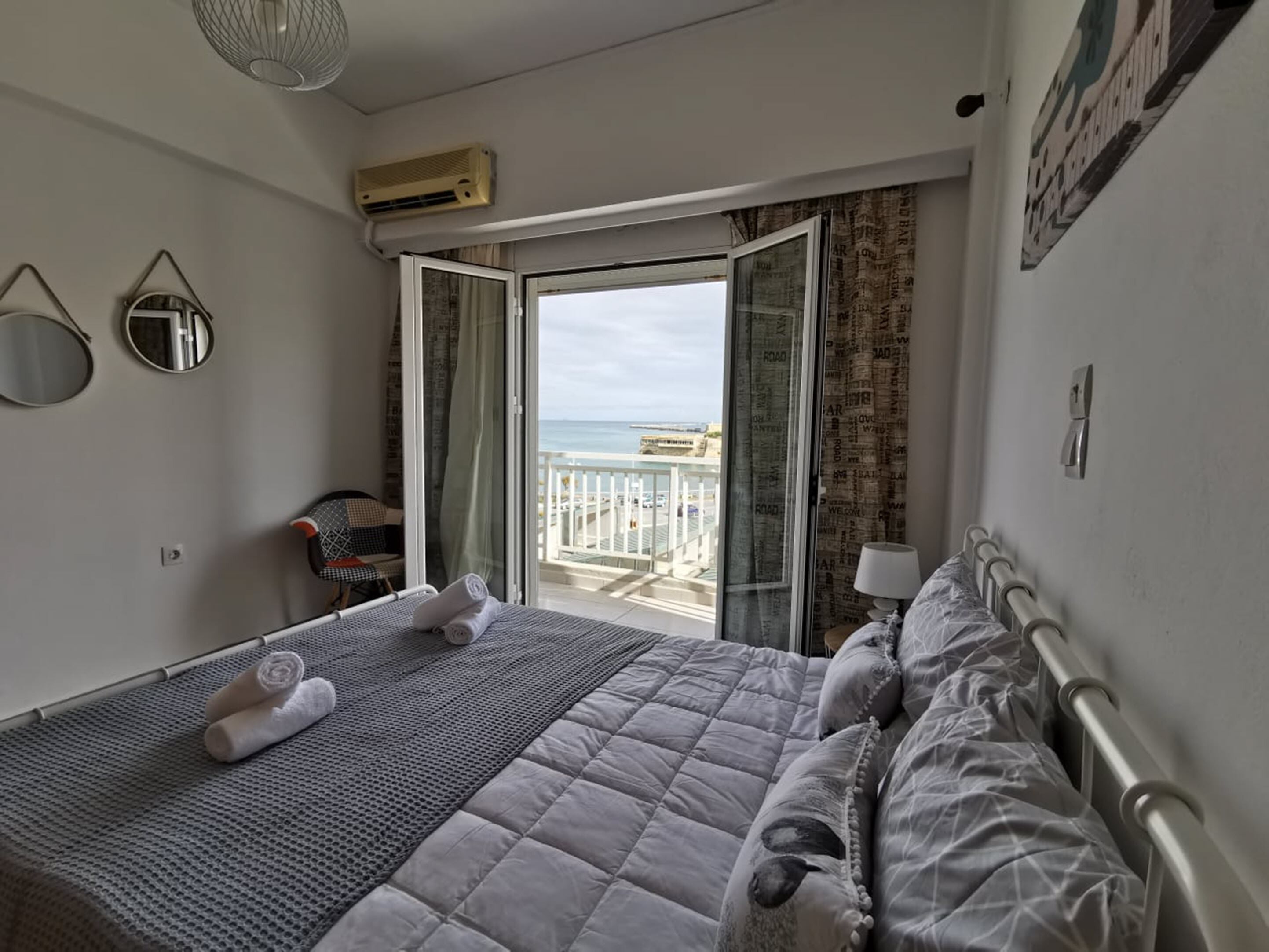 A hotel room with a bed, a chair, a balcony with a view of the sea, and an air conditioning unit.