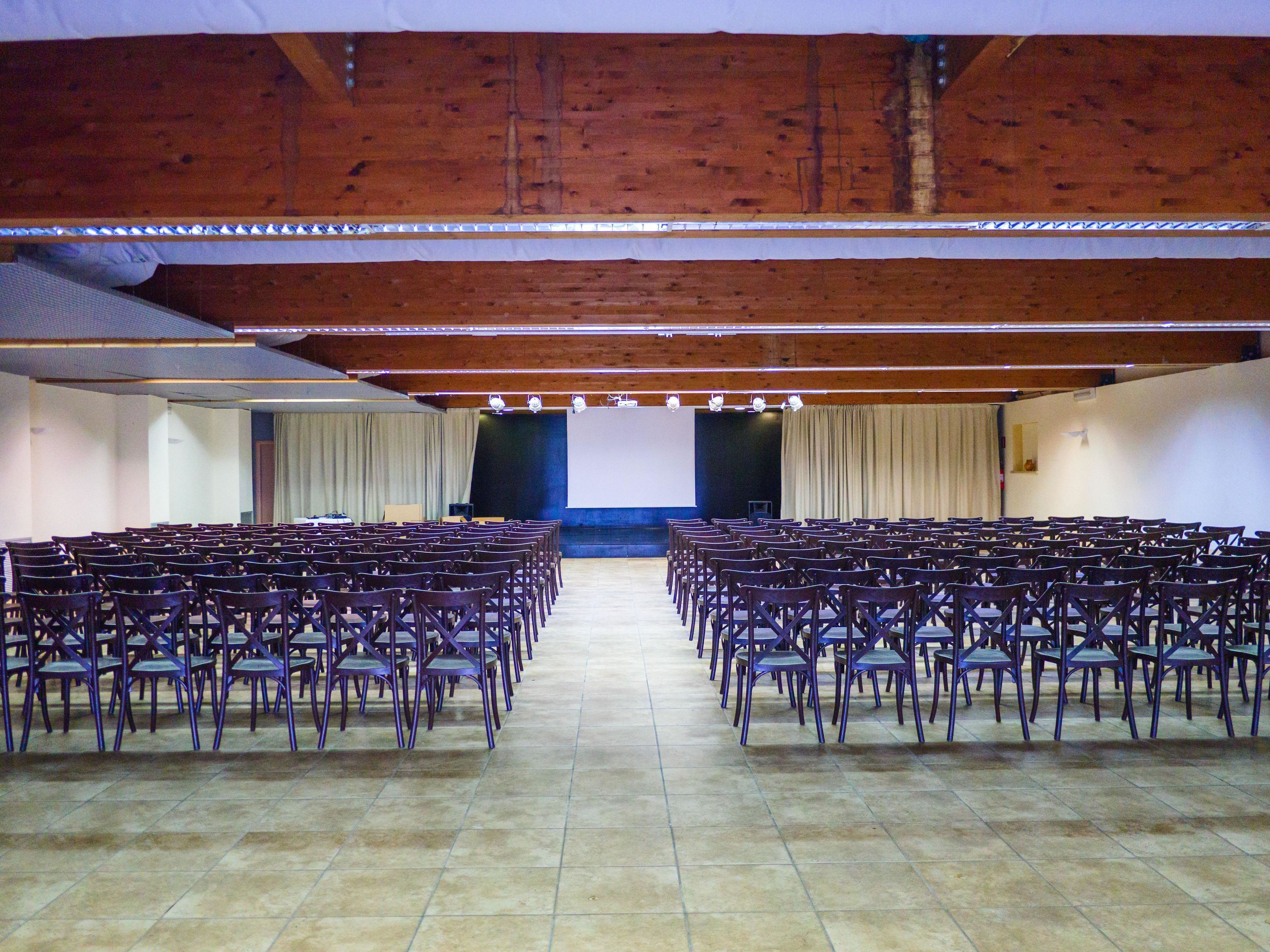 A conference room with rows of chairs, a stage with a screen, and wooden ceiling beams.