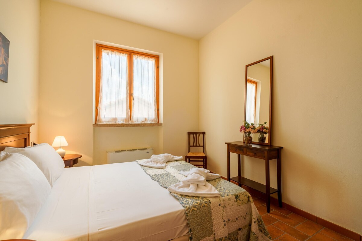 Een hotelkamer met een bed, een raam met gordijnen, een stoel, een tafel met een vaas en een spiegel.