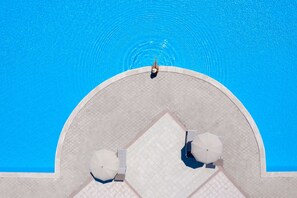 Luchtbeeld van een zwembad met betegeld zonnedek, twee parasols en een persoon aan de rand.