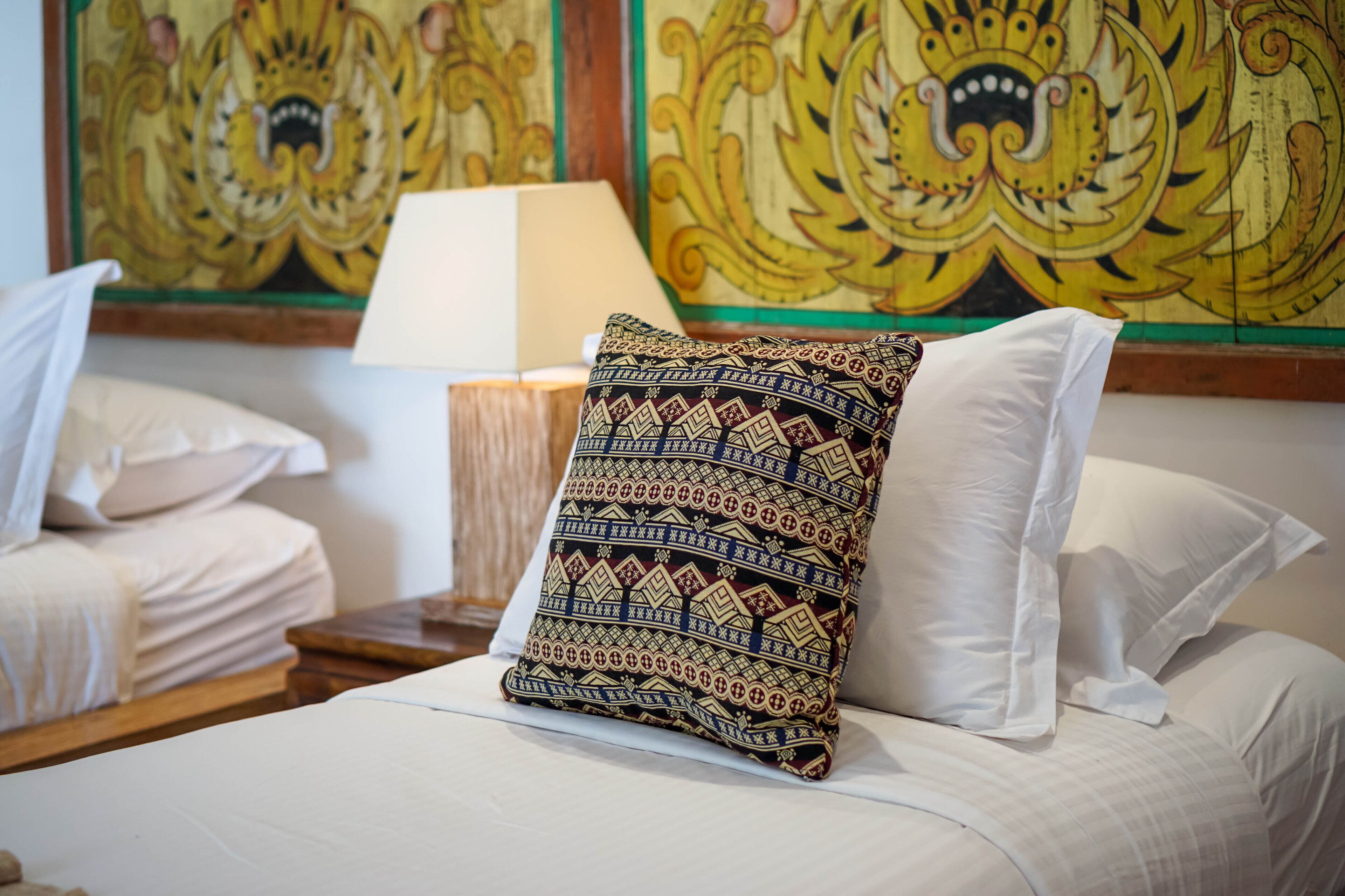 A neatly made bed with patterned pillows, a bedside lamp, and a wall with traditional artwork.