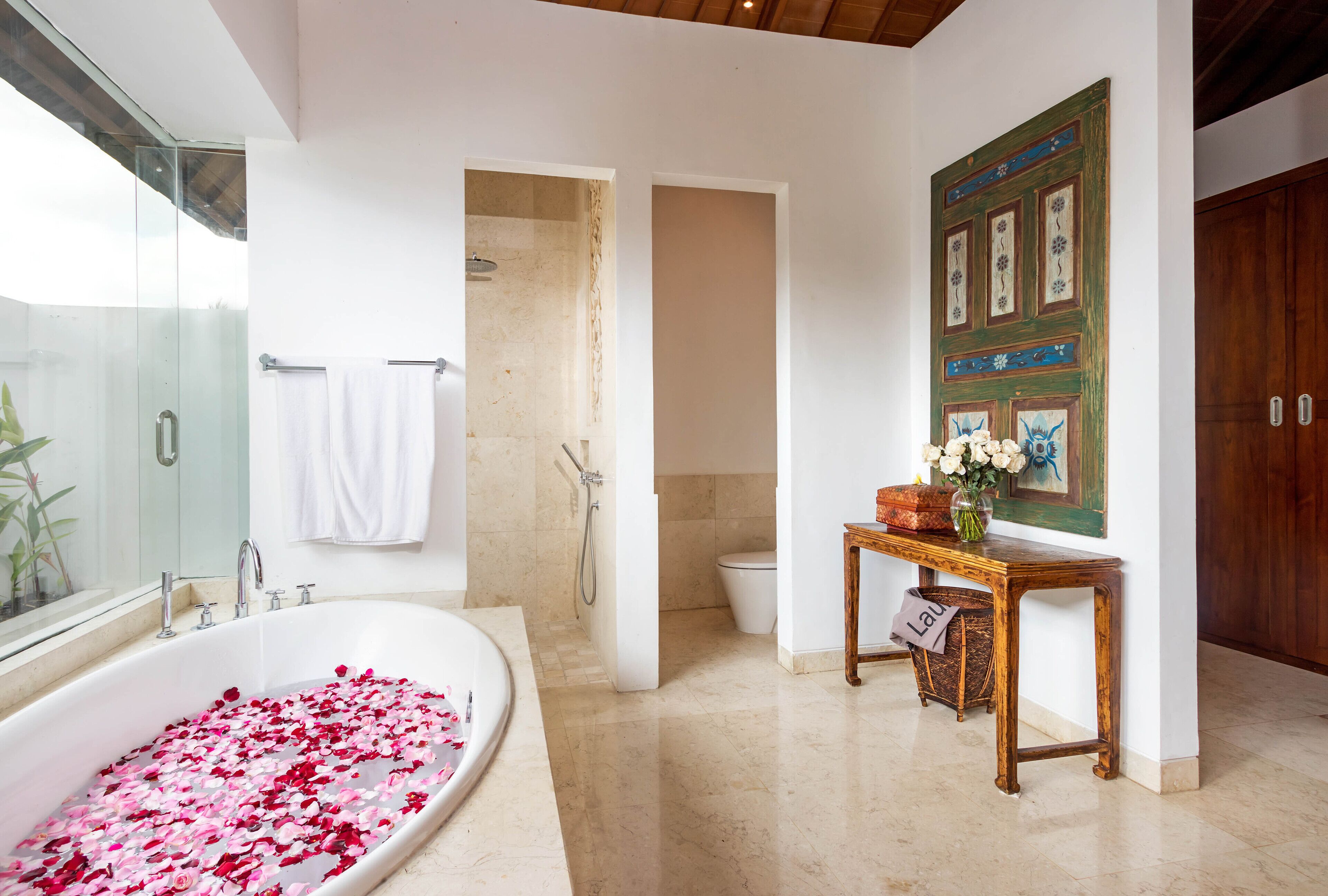 A modern bathroom with a large bathtub filled with rose petals, a glass shower enclosure, and a wooden table with a vase of flowers.
