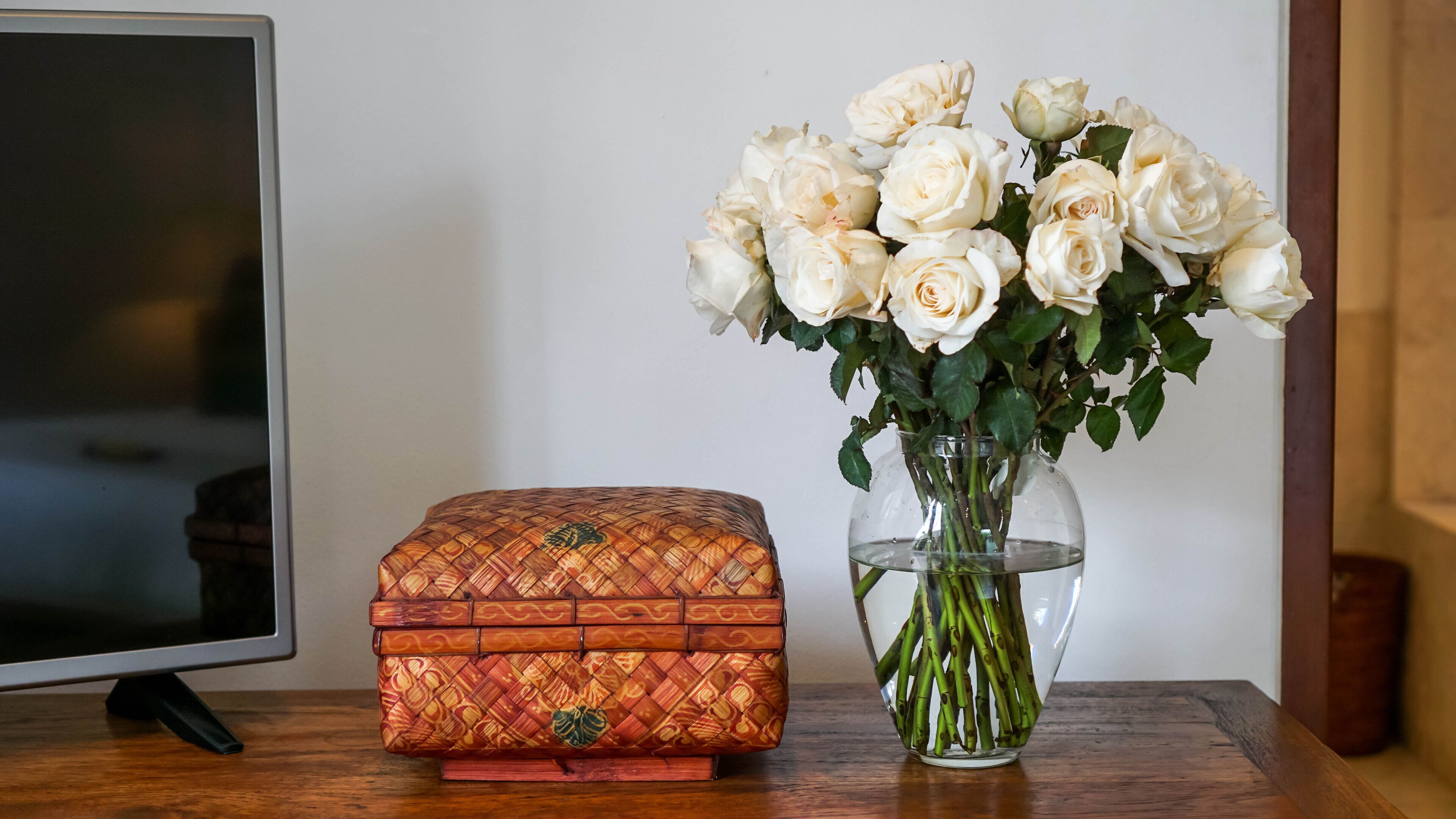 Un cofre de madera con un diseño tejido y un jarrón con rosas blancas sobre una mesa de madera.