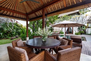 A covered outdoor seating area with wicker furniture and a central table, surrounded by lush greenery and a pool.