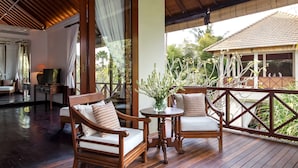 A wooden deck with two cushioned chairs and a small table, overlooking a garden and a traditional building. - Villa Rubina (Seminyak)