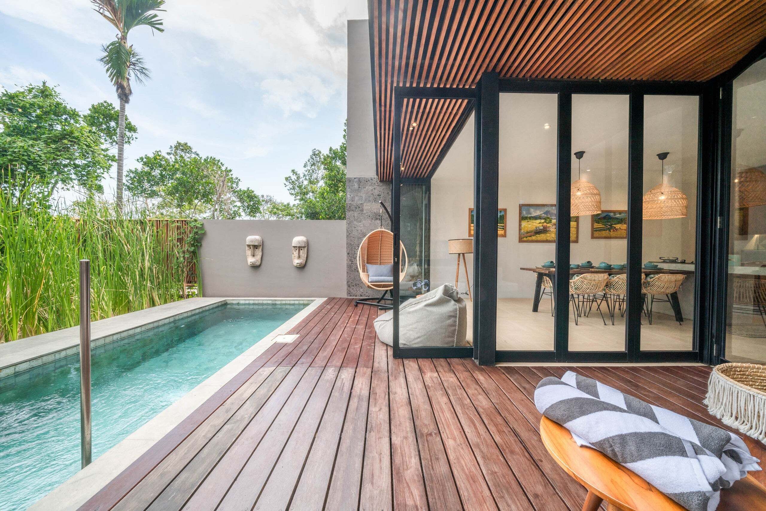 A modern outdoor pool area with a wooden deck, a hanging chair, and a glass door leading to a dining area with a table and chairs.