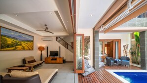 A modern living room with a large painting, a sofa, a TV, and a wooden floor leading to a pool area. - Villa Seacluded 1 (Seminyak)