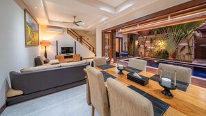 A modern living room with a sofa, dining table, and chairs, a staircase, and a pool area with plants. - Villa Seacluded 1 (Seminyak)