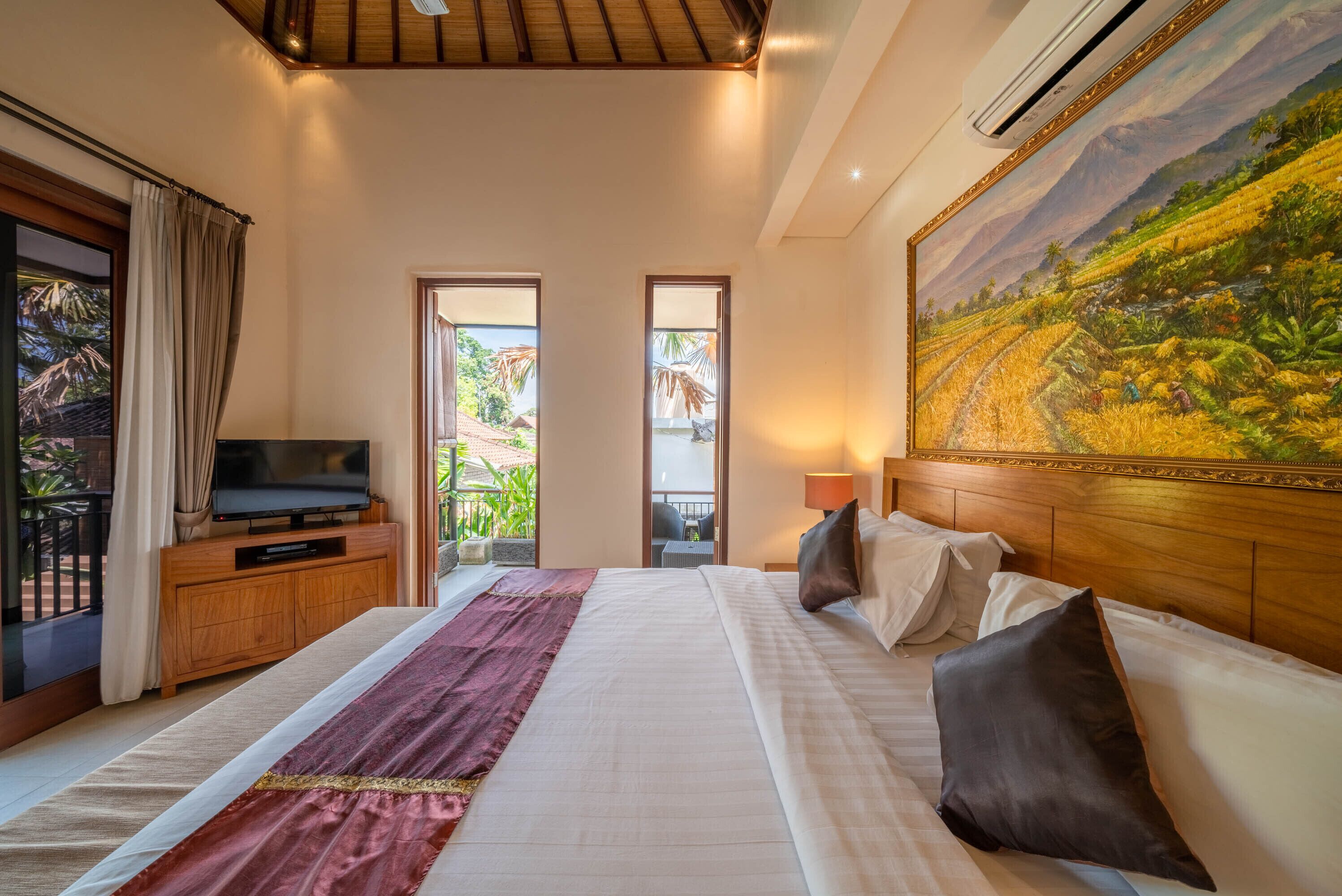 A hotel room with a large bed, a television on a wooden cabinet, a painting of a landscape, and a balcony with curtains.