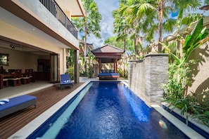 Una piscina con sillas de descanso, un área de estar techada y plantas tropicales.