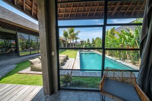 A modern pool area with a clear view through large windows, a wooden deck, and outdoor seating. - Villa Wirtiga (Canggu)