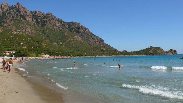 Une plage aux eaux cristallines, avec des montagnes rocheuses en arrière-plan, et des gens qui se baignent.