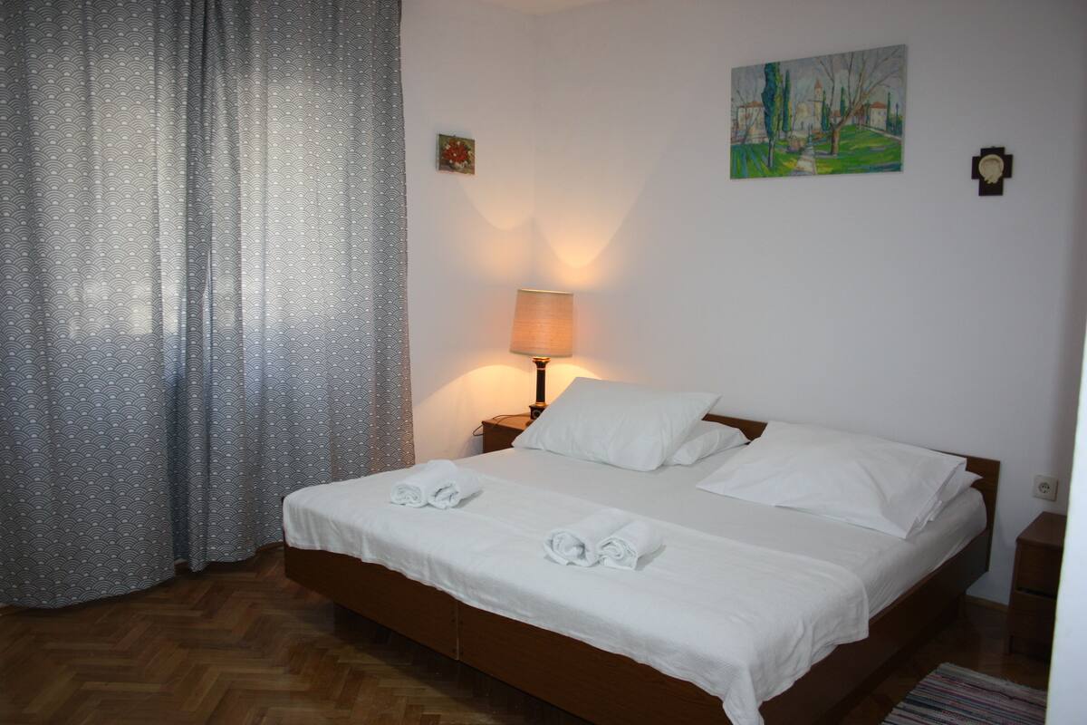 A neatly made bed with white linens and towels, a lamp on the nightstand, and a painting on the wall.
