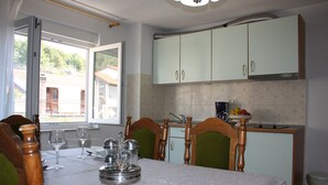 A dining area with a table set for four, a window with a view of buildings, and a kitchen with light green cabinets. - Relax Adriatic House (Omis)