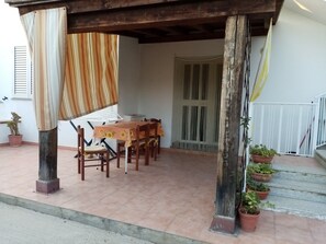 Un área de comedor al aire libre techada con una mesa y sillas de madera, plantas en macetas y piso de baldosas.
