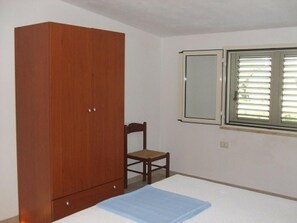 A bedroom with a wooden wardrobe, a single bed, a chair, and a window with blinds.