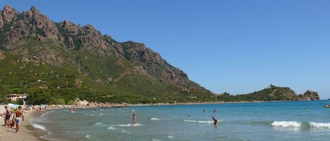 Una playa con aguas cristalinas, gente nadando y montañas al fondo.