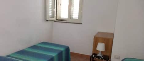 Una habitación pequeña y ordenada con dos camas individuales, una mesita de noche de madera con lámpara y una ventana con persianas.