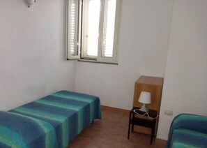 Una habitación pequeña y ordenada con dos camas individuales, una mesita de noche de madera con lámpara y una ventana con persianas.