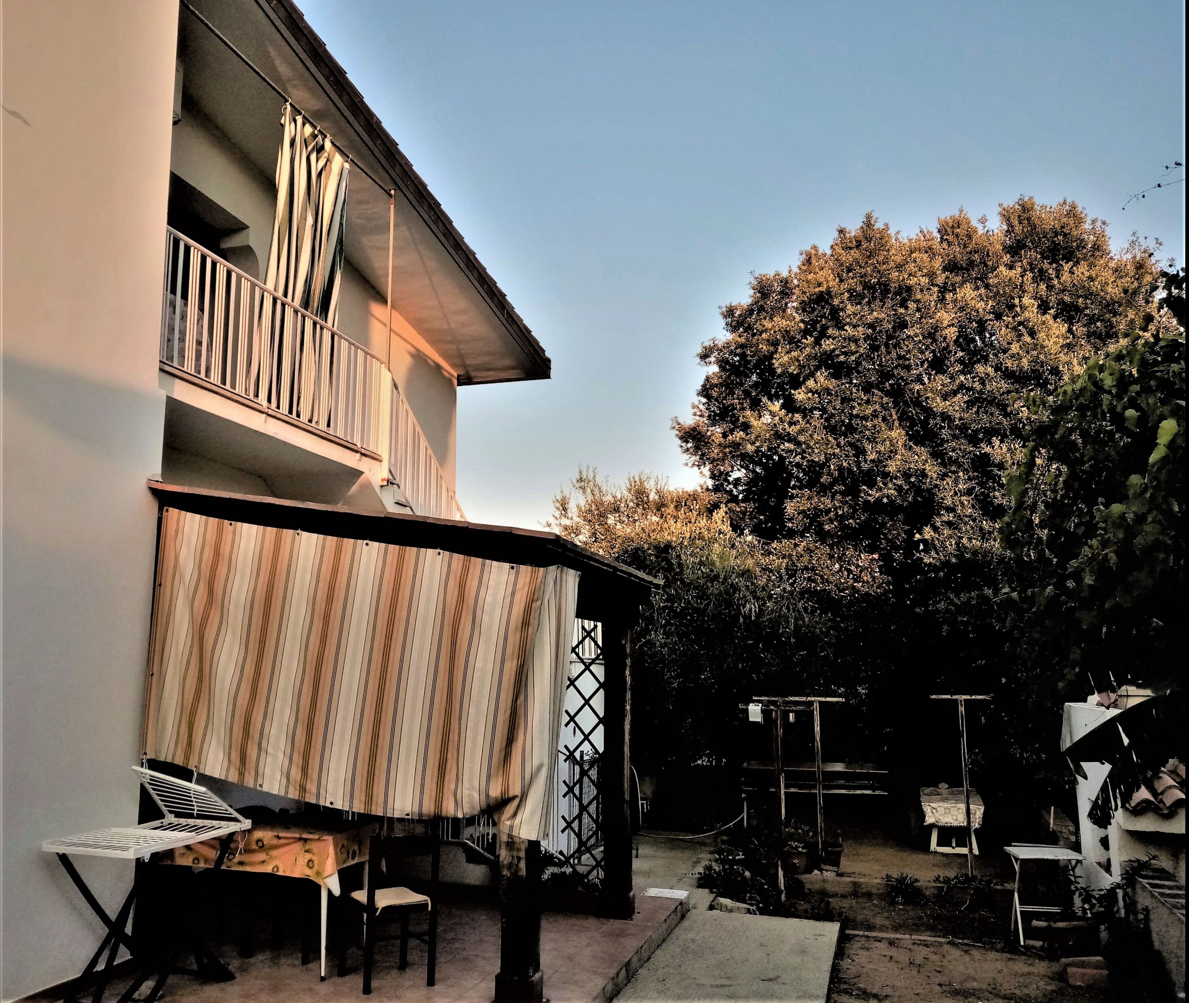 Un bâtiment à deux étages doté d’un balcon, d’un espace extérieur couvert aménagé en coin salon et d’un jardin avec des arbres et des plantes.