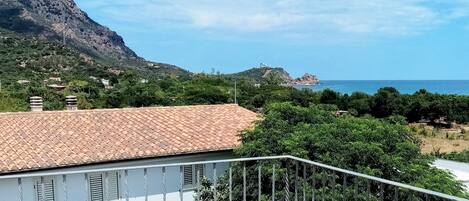 Un balcón con mantel floreado amarillo, una silla blanca y vista a un paisaje costero con montañas y el mar.