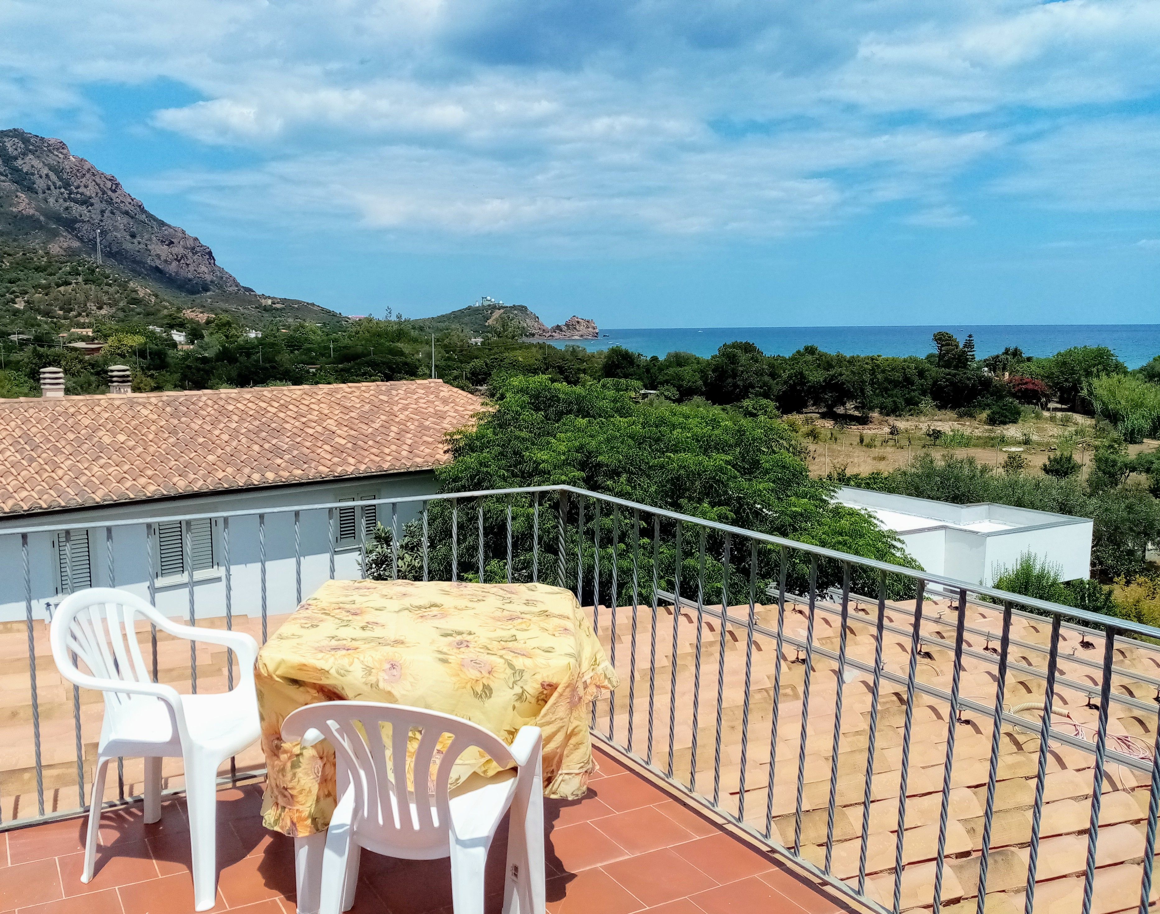 Ein Balkon mit Tisch und zwei Stühlen, mit Blick auf eine Küstenlandschaft mit Berg und Vegetation.