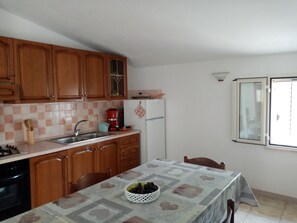 A kitchen with wooden cabinets, a sink, a stove, and a refrigerator. There is a table with a tablecloth and a basket of fruit. - Always on Foot 3 min to the Beach Center With Double Terrace (Tertenia)
