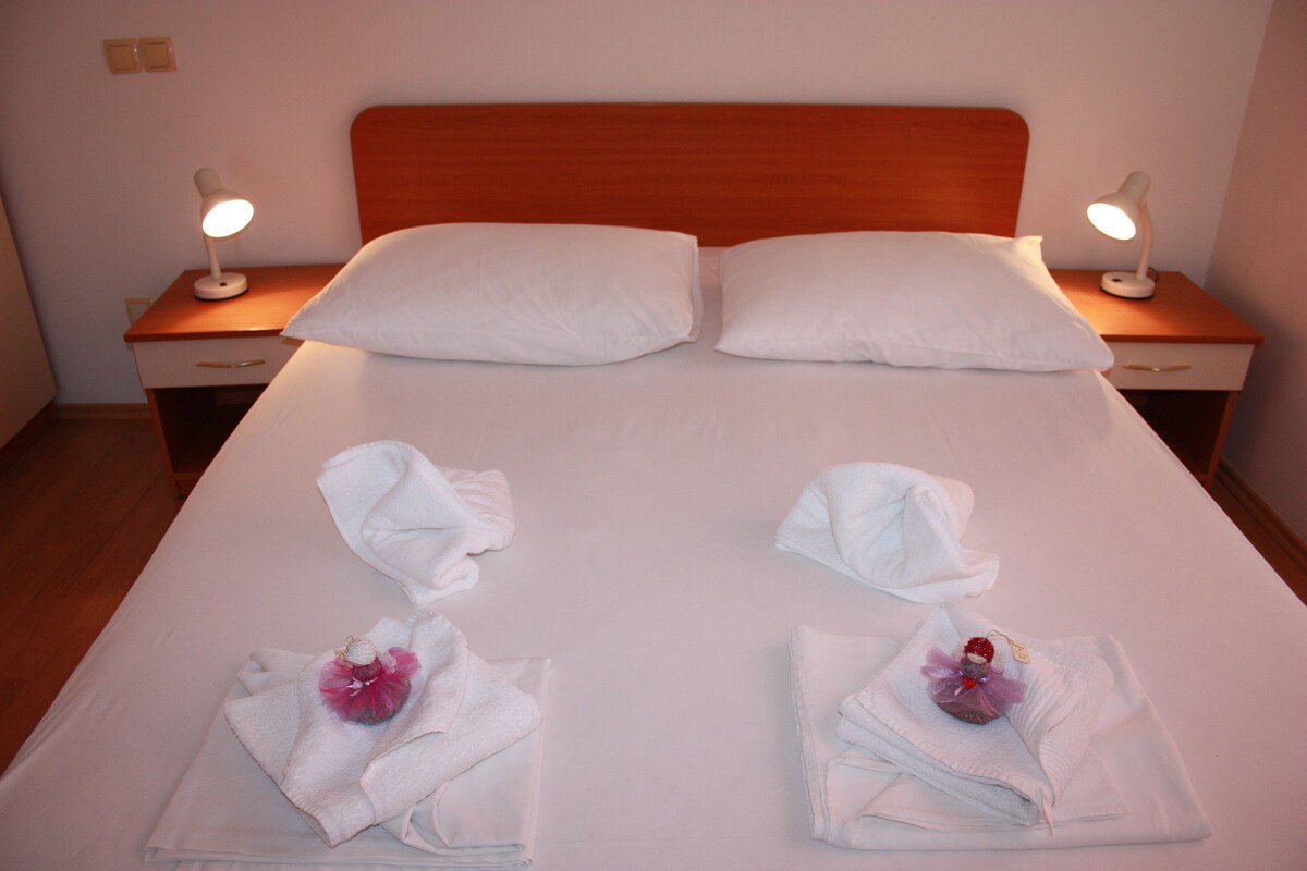 A neatly made bed with two pillows, two bedside tables with lamps, and towels with flowers on the bed.