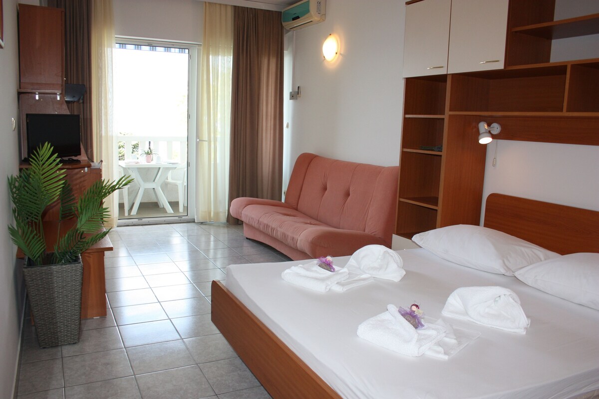 A hotel room with a bed, a sofa, a TV, a potted plant, and a balcony with a table and chairs.
