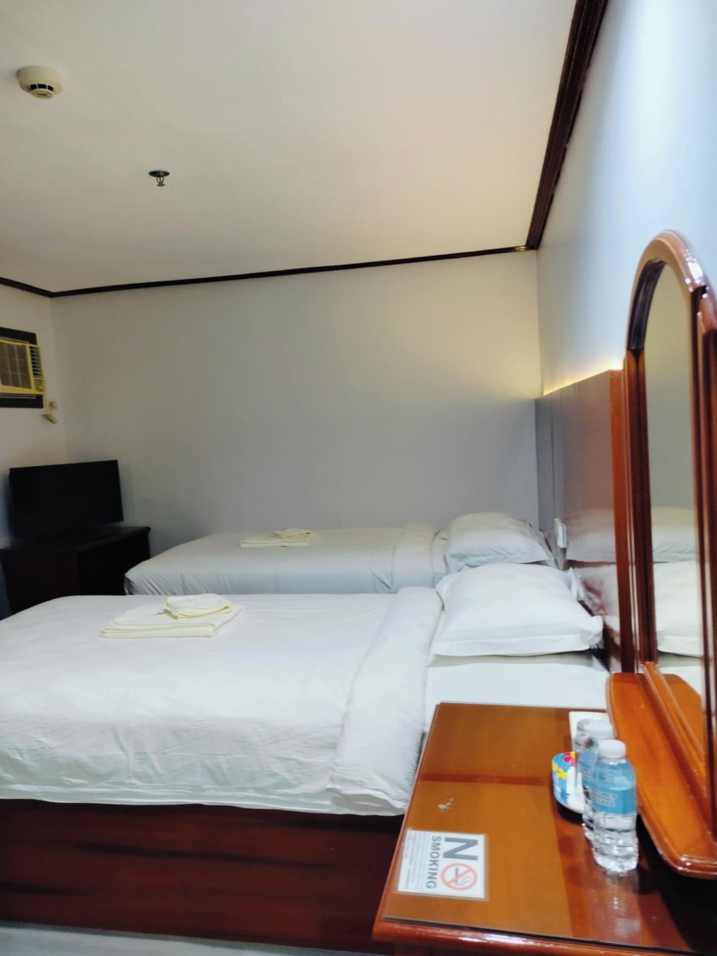 Habitación de hotel con dos camas, un tocador de madera, un espejo, un cartel de 'No Fumar' y una botella de agua.