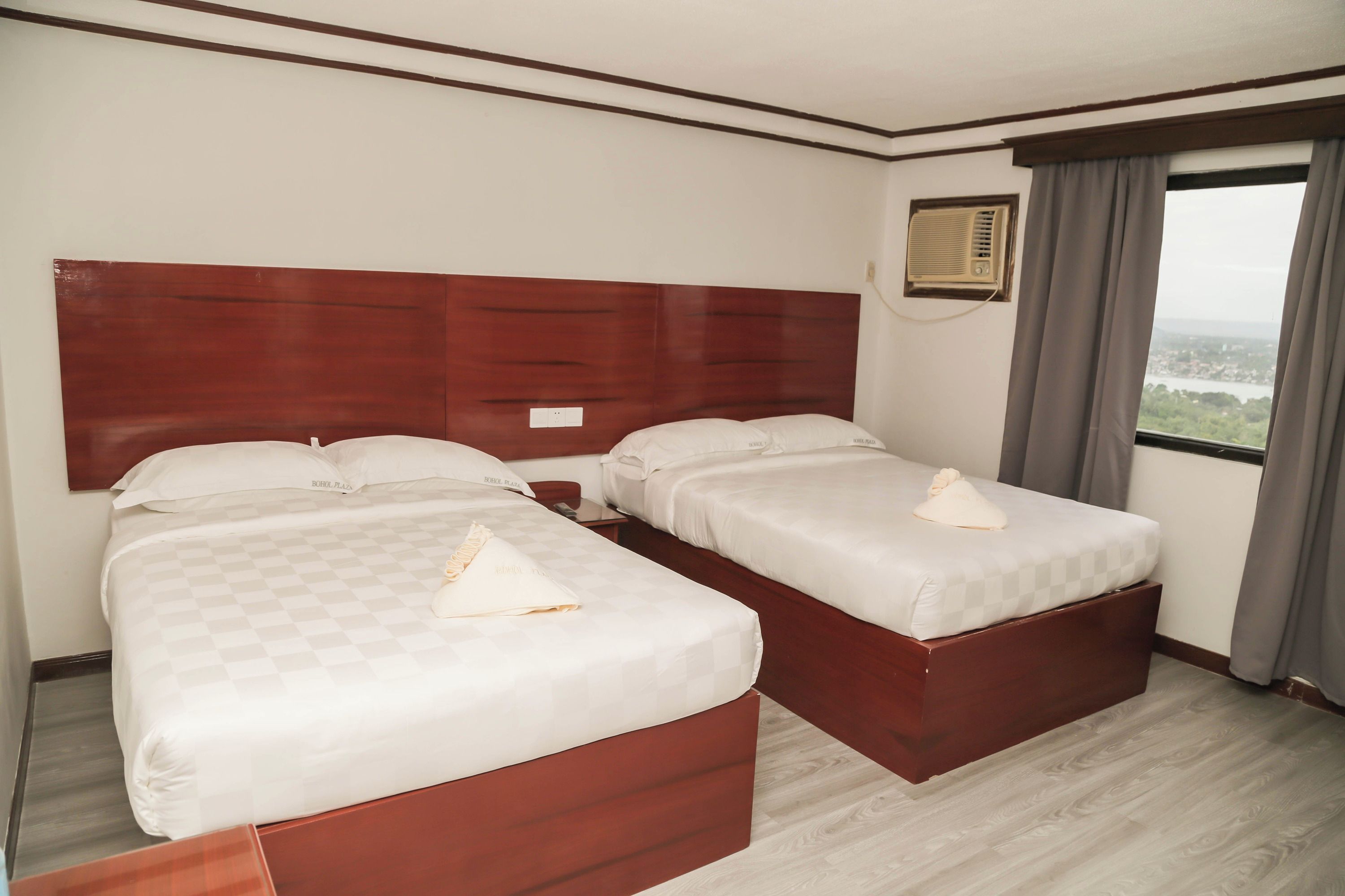 A hotel room with two beds, each with a wooden headboard and white bedding. There is a window with curtains, an air conditioning unit on the wall, and a view of the cityscape.